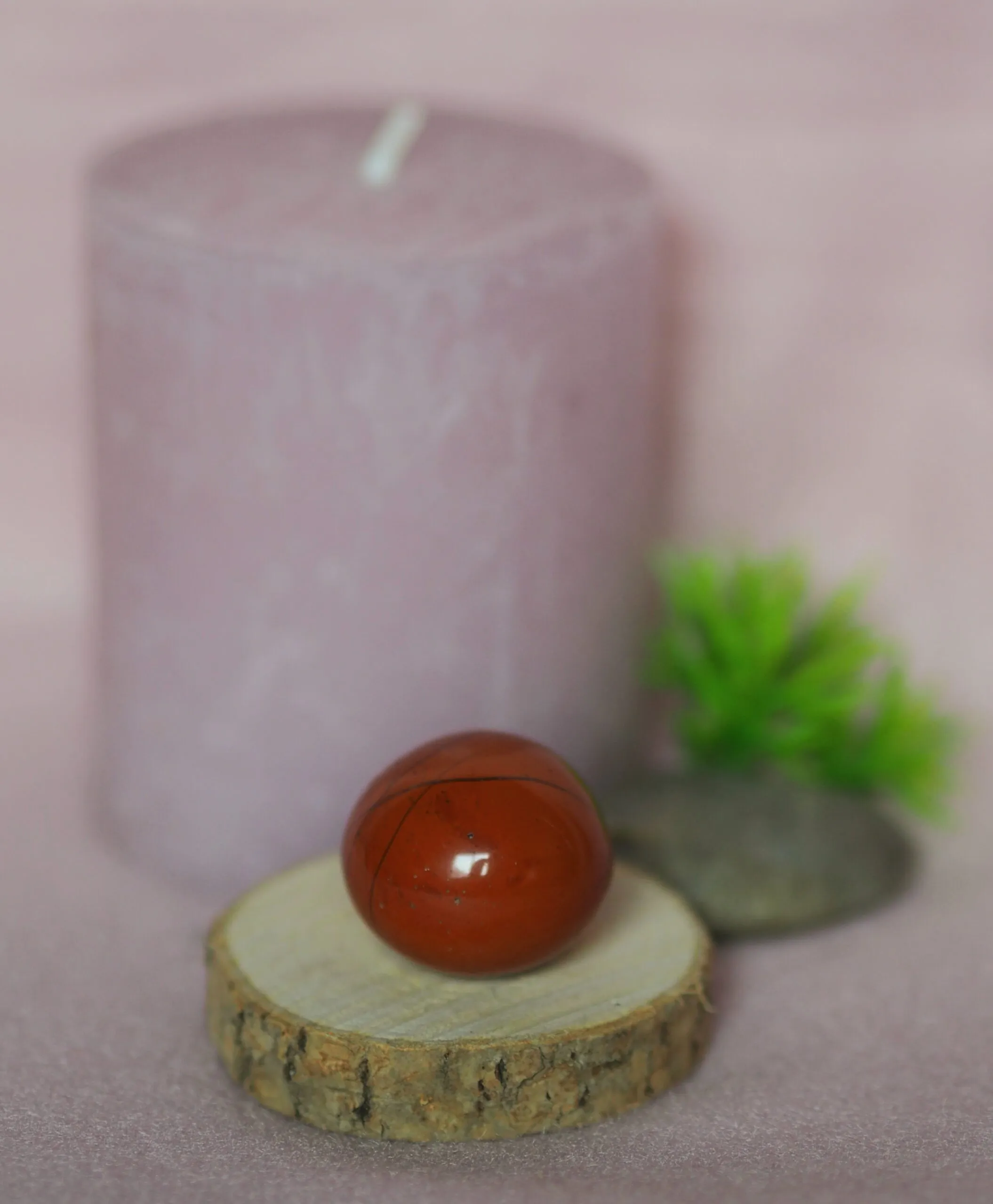 Pierre roulée de jaspe rouge, polie et brillante, posée sur une rondelle de bois devant une bougie rose