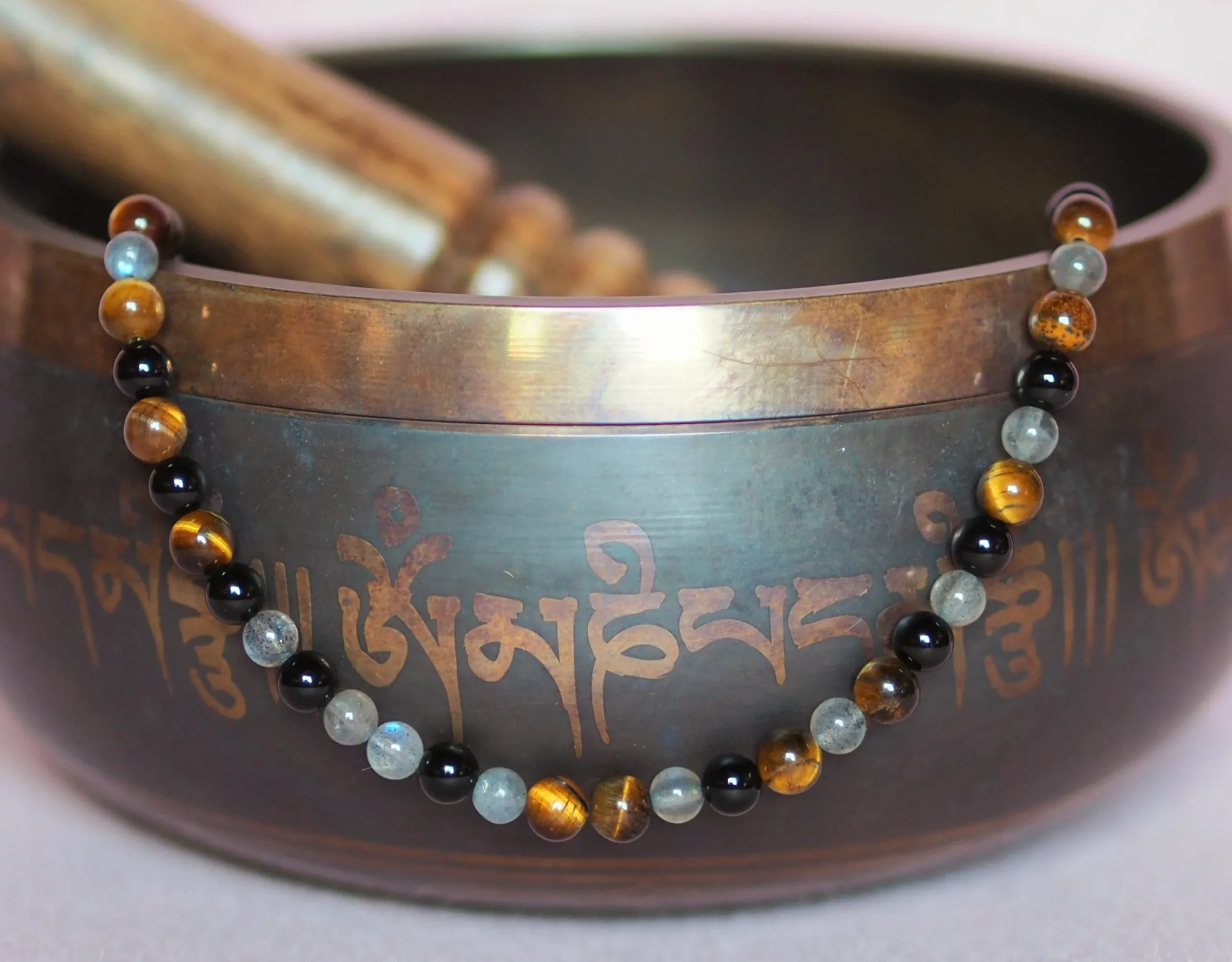Collier de perles d’œil de tigre, d’obsidienne noire et de labradorite, disposé sur le bord d’un bol chantant gravé