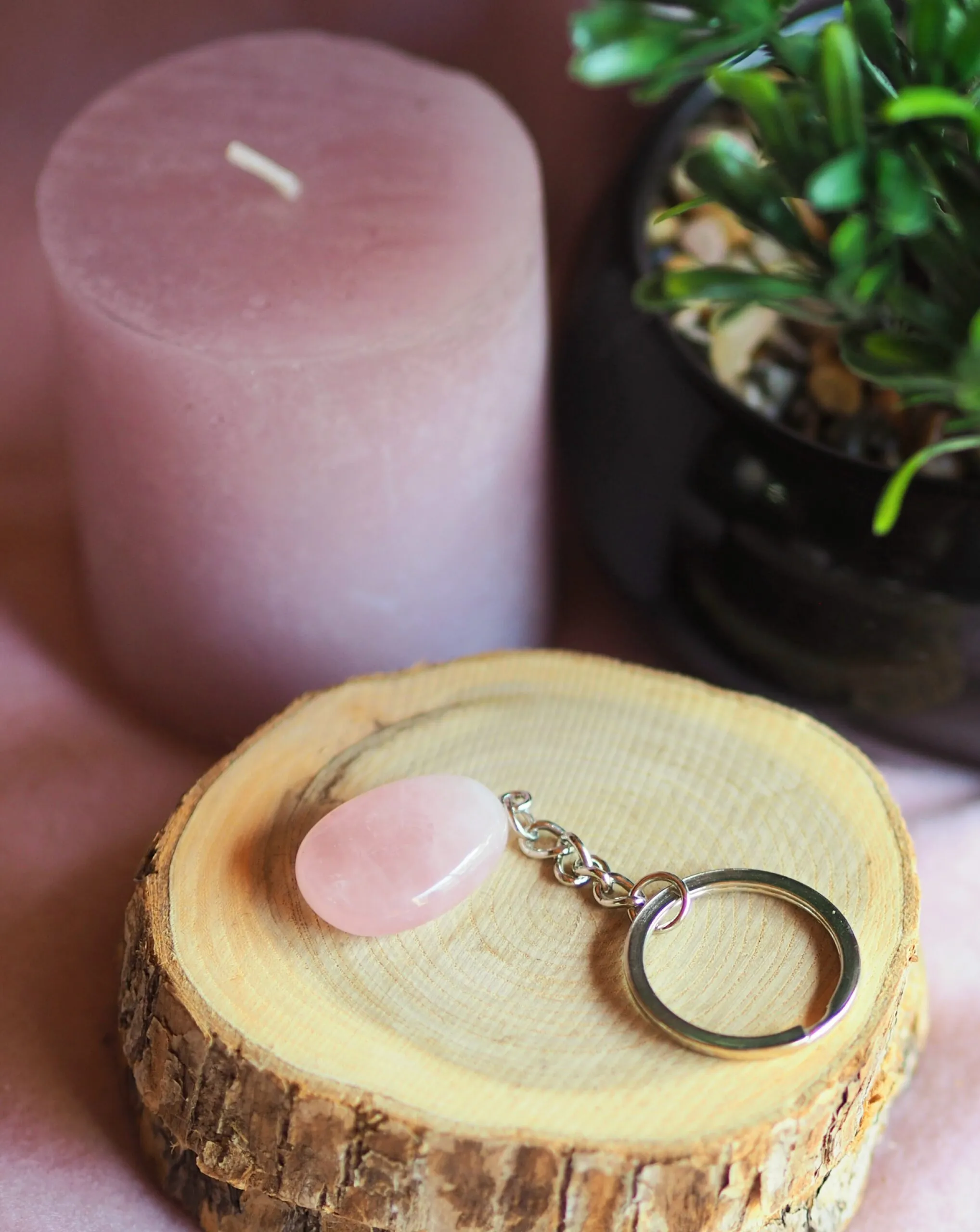 Porte-clés en quartz rose pâle, pierre plate sur une chaînette et un anneau argentés, posé sur une rondelle de bois près d’une bougie