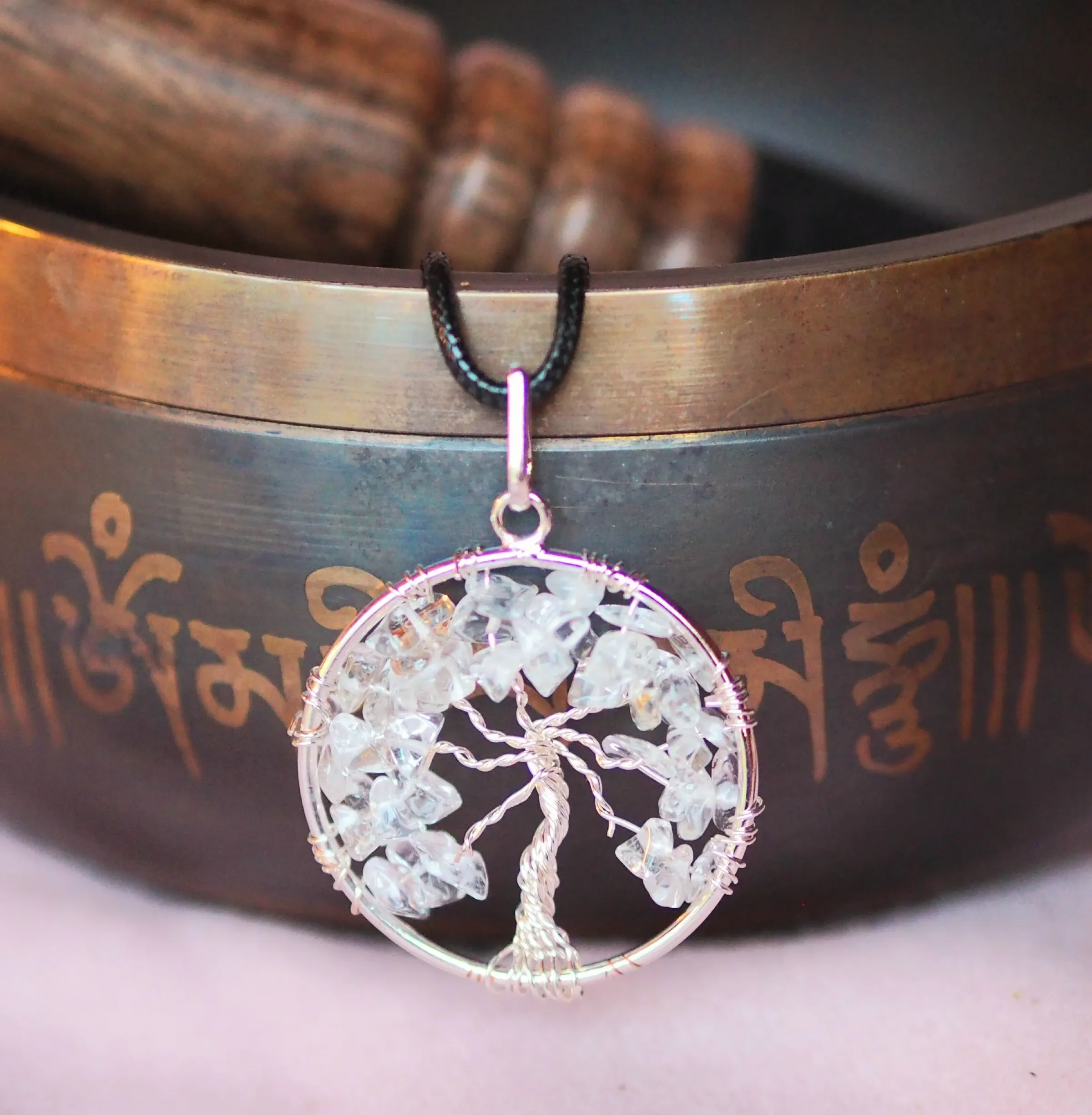 Pendentif rond en fil argenté formant un arbre de vie aux branches garnies d’éclats de cristal de roche, sur un cordon noir, devant un bol chantant