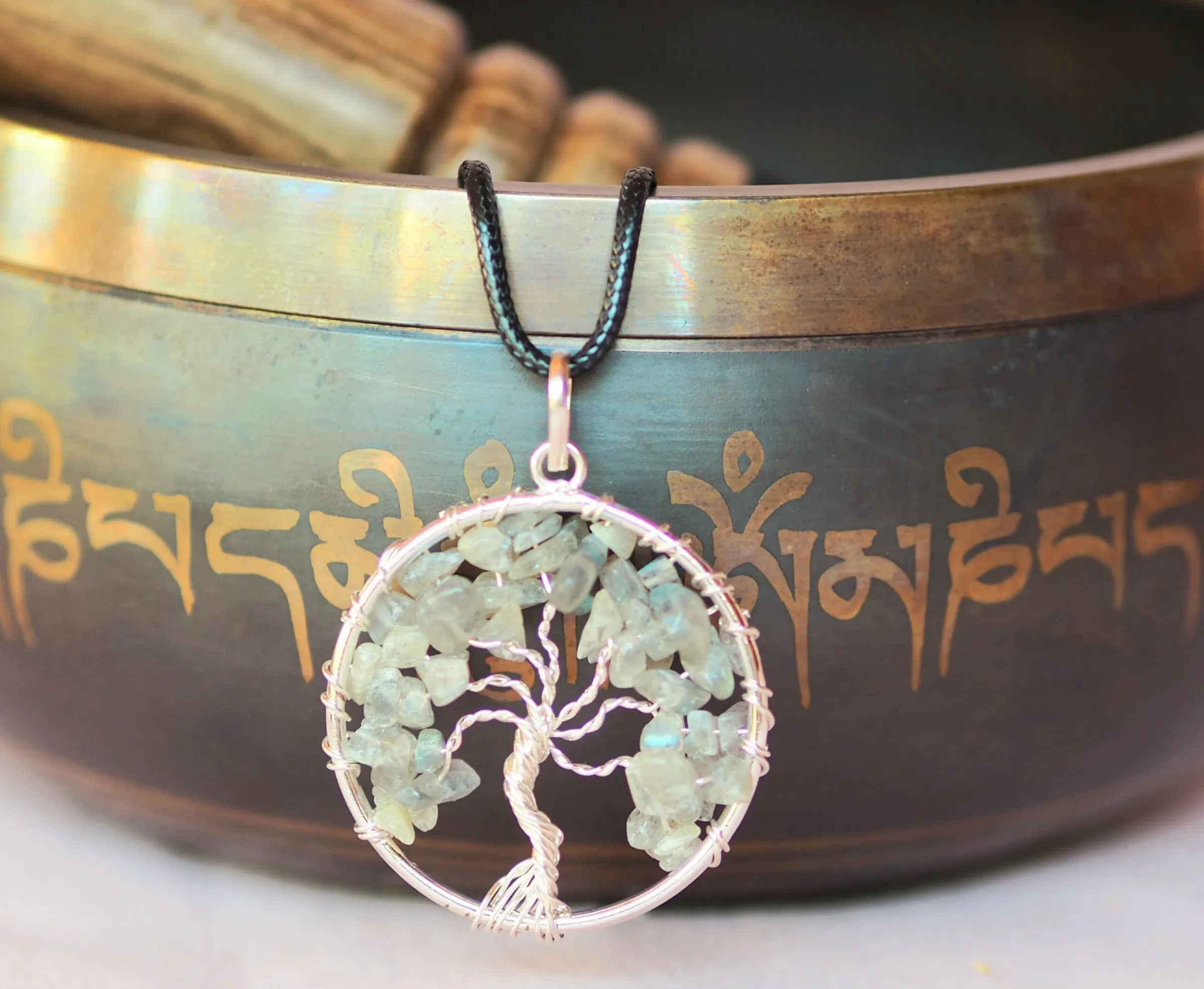 Pendentif rond en fil argenté formant un arbre de vie aux branches garnies d’éclats de labradorite grise aux reflets irisés, sur un cordon noir, devant un bol chantant
