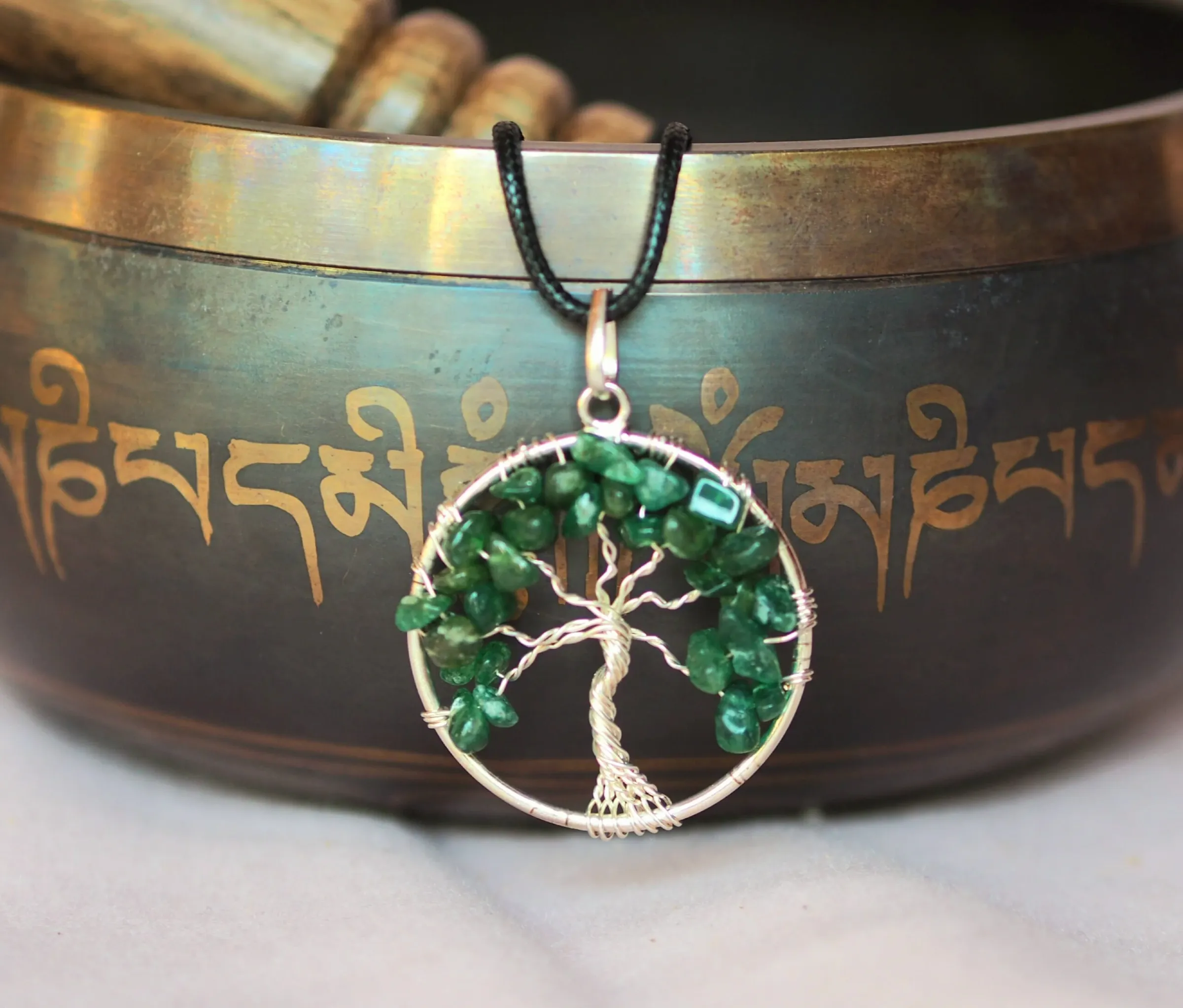 Pendentif rond en fil argenté formant un arbre de vie aux branches garnies d’éclats d’aventurine vert foncé, sur un cordon noir, devant un bol chantant