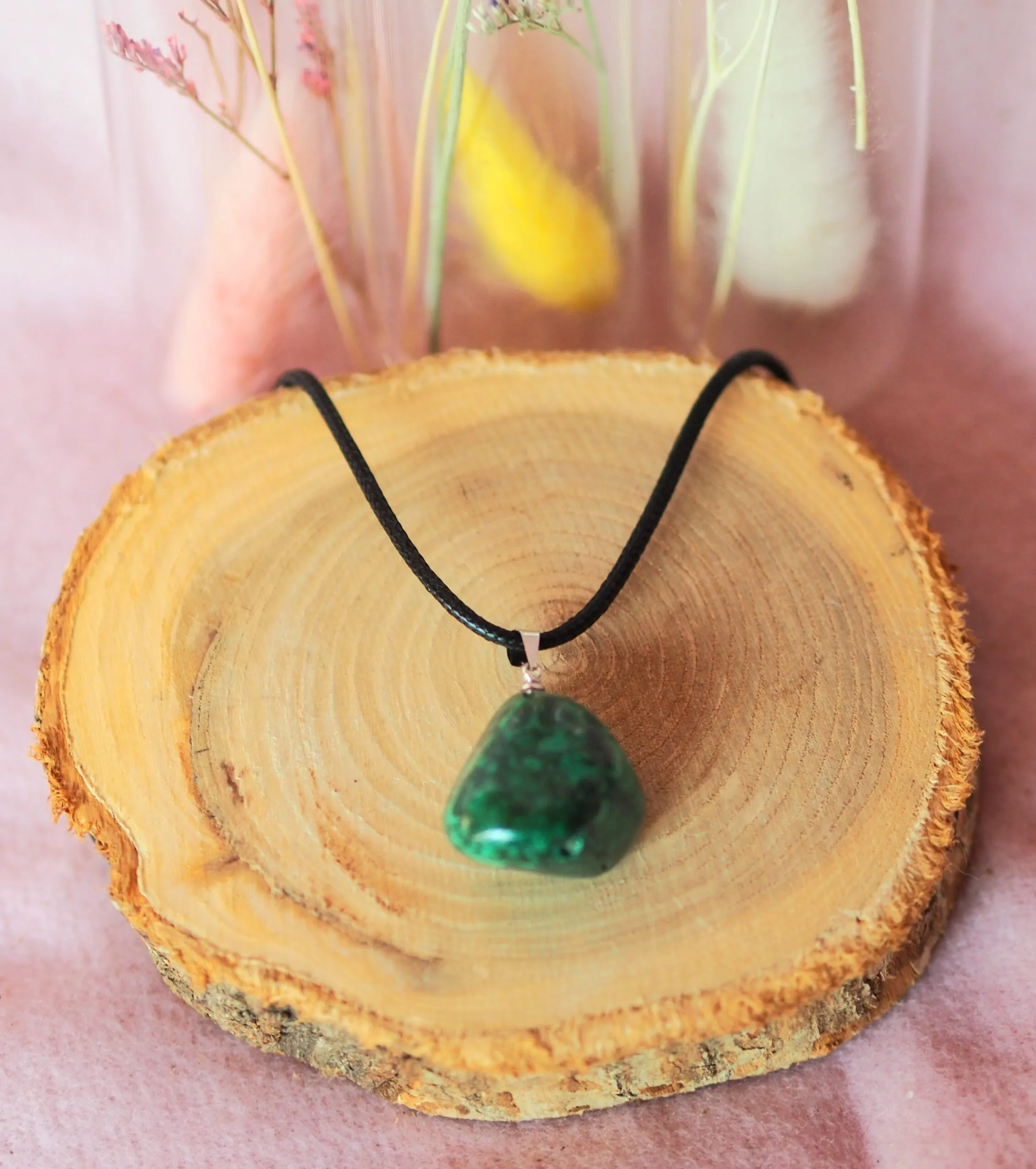 Pendentif en pierre roulée vert profond, avec une bélière argentée sur un cordon de cuir noir, posé sur une rondelle de bois