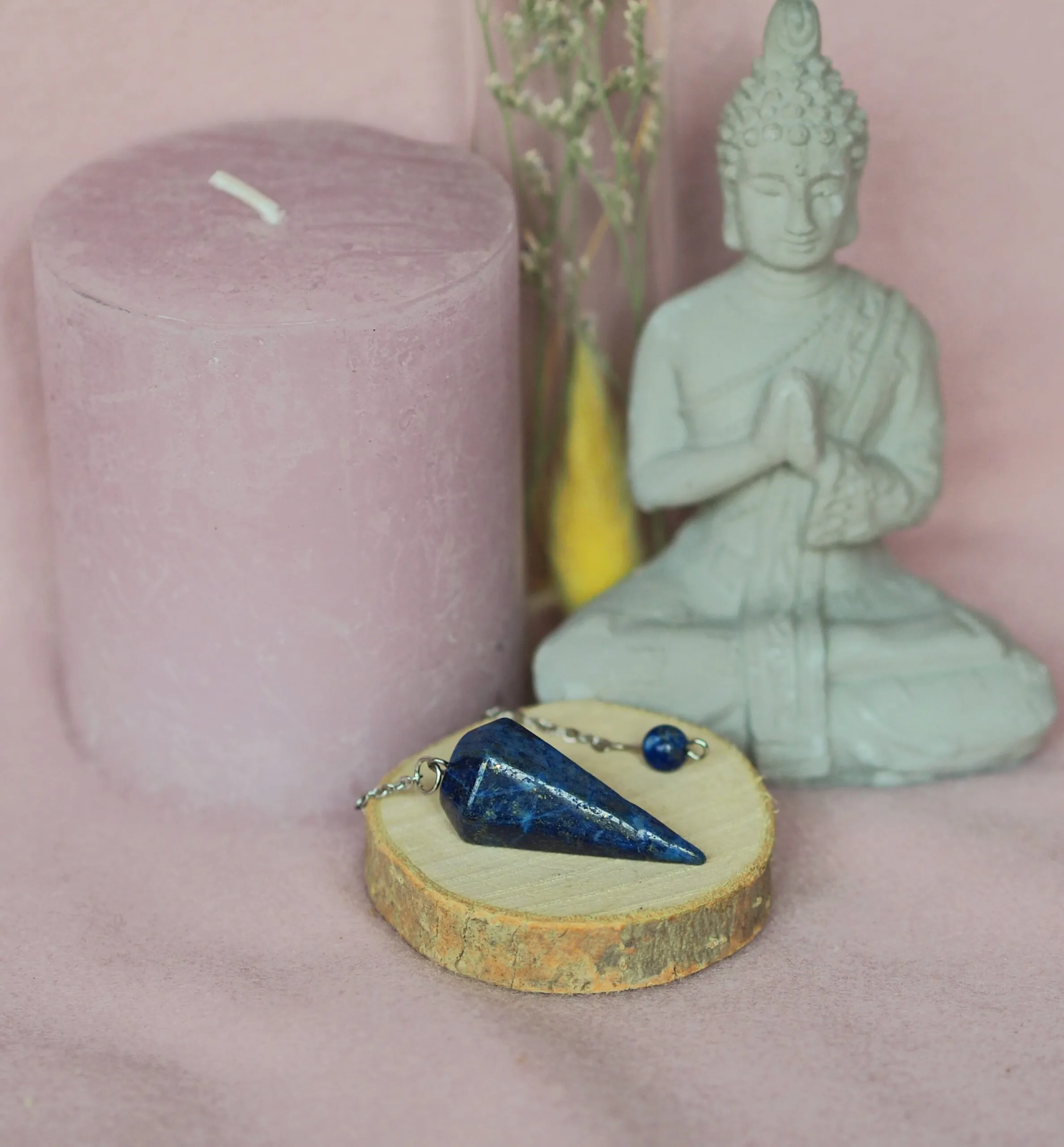 Pendule conique facetté en lapis-lazuli bleu nuit, posé avec sa chaînette sur une rondelle de bois, devant une statuette de Bouddha