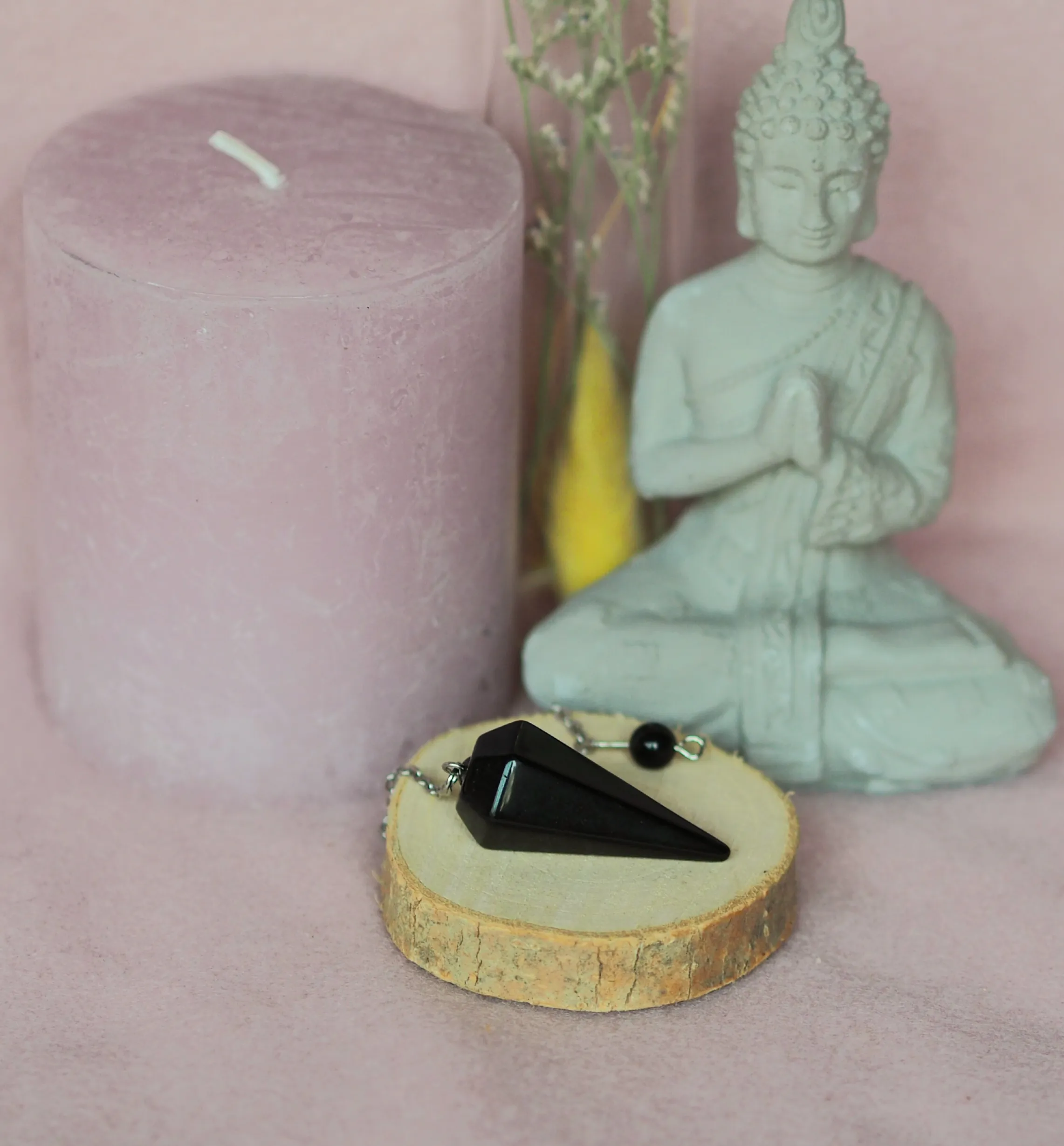 Pendule conique facetté en obsidienne noire brillante, posé avec sa chaînette sur une rondelle de bois devant une statuette de Bouddha