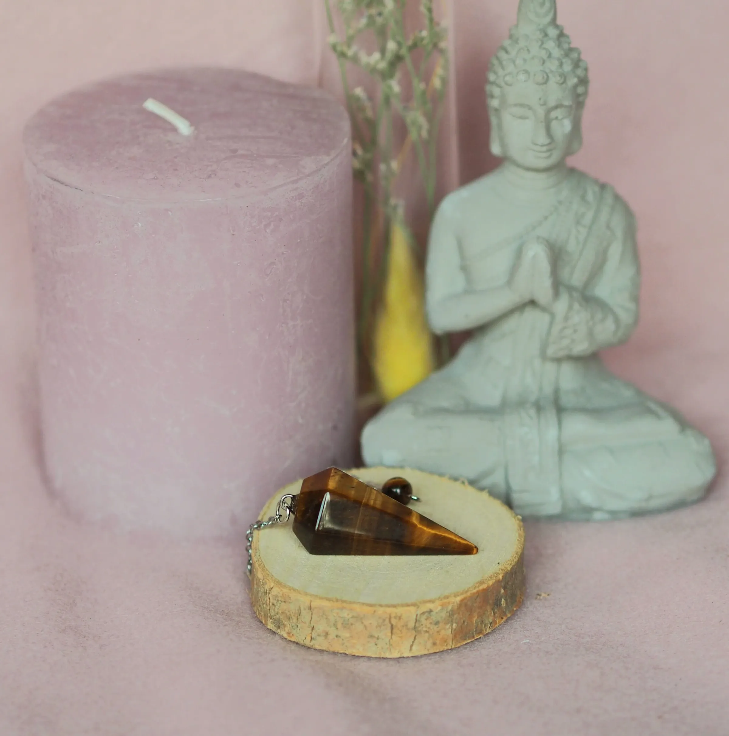 Pendule conique facetté en œil de tigre brun doré, posé avec sa chaînette sur une rondelle de bois devant une statuette de Bouddha