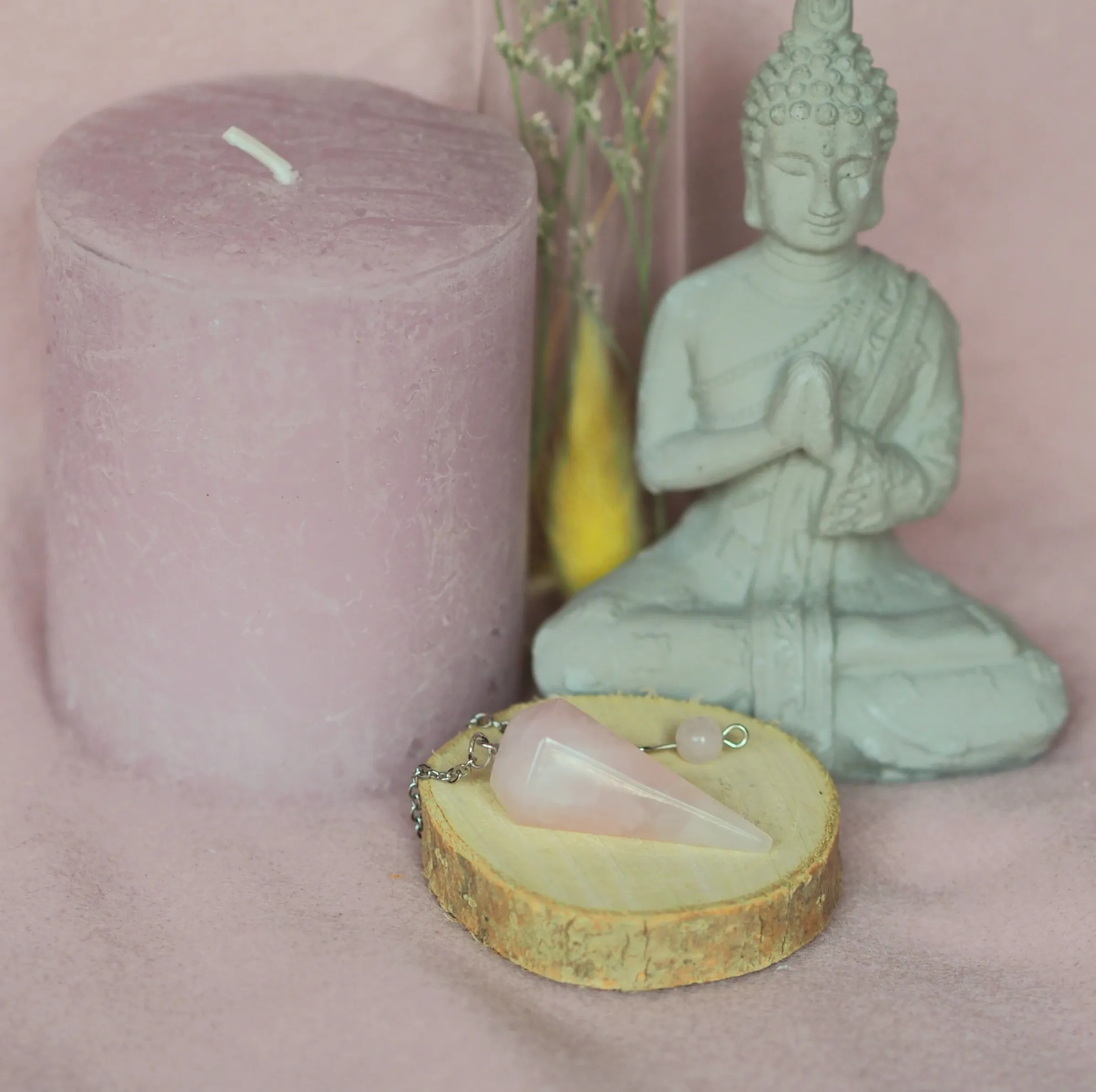 Pendule conique en quartz rose pâle, posé avec sa chaînette sur une rondelle de bois devant une statuette de Bouddha