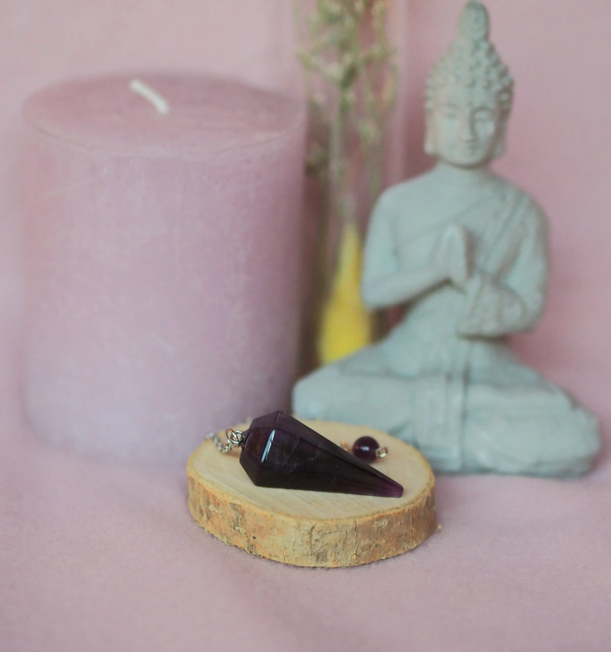 Pendule conique facetté en améthyste violette, posé avec sa chaînette sur une rondelle de bois, devant une bougie rose et une statuette de Bouddha