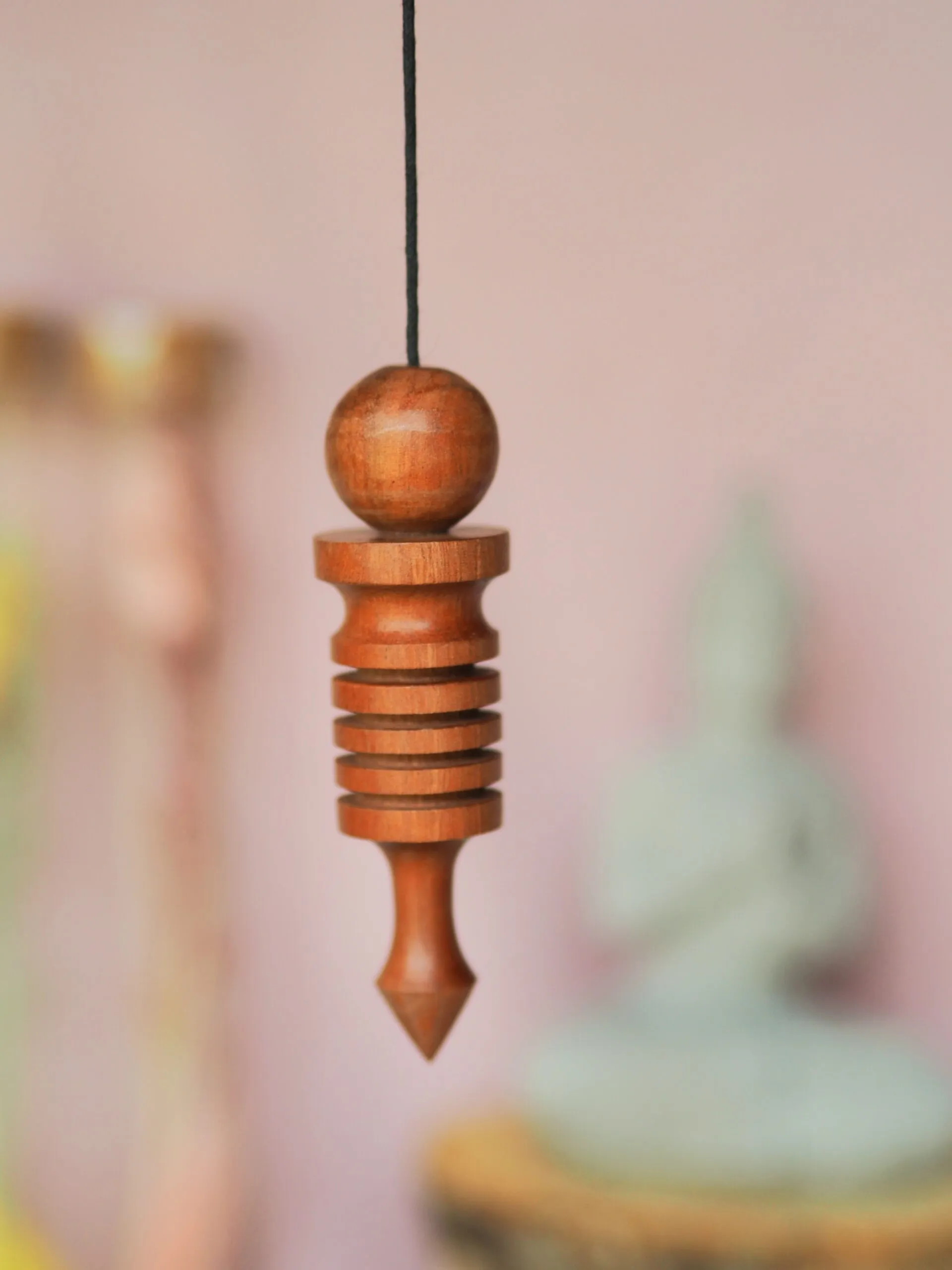 Pendule Isis en bois clair, une sphère au-dessus de disques superposés et d’une pointe, suspendu devant un mur rose