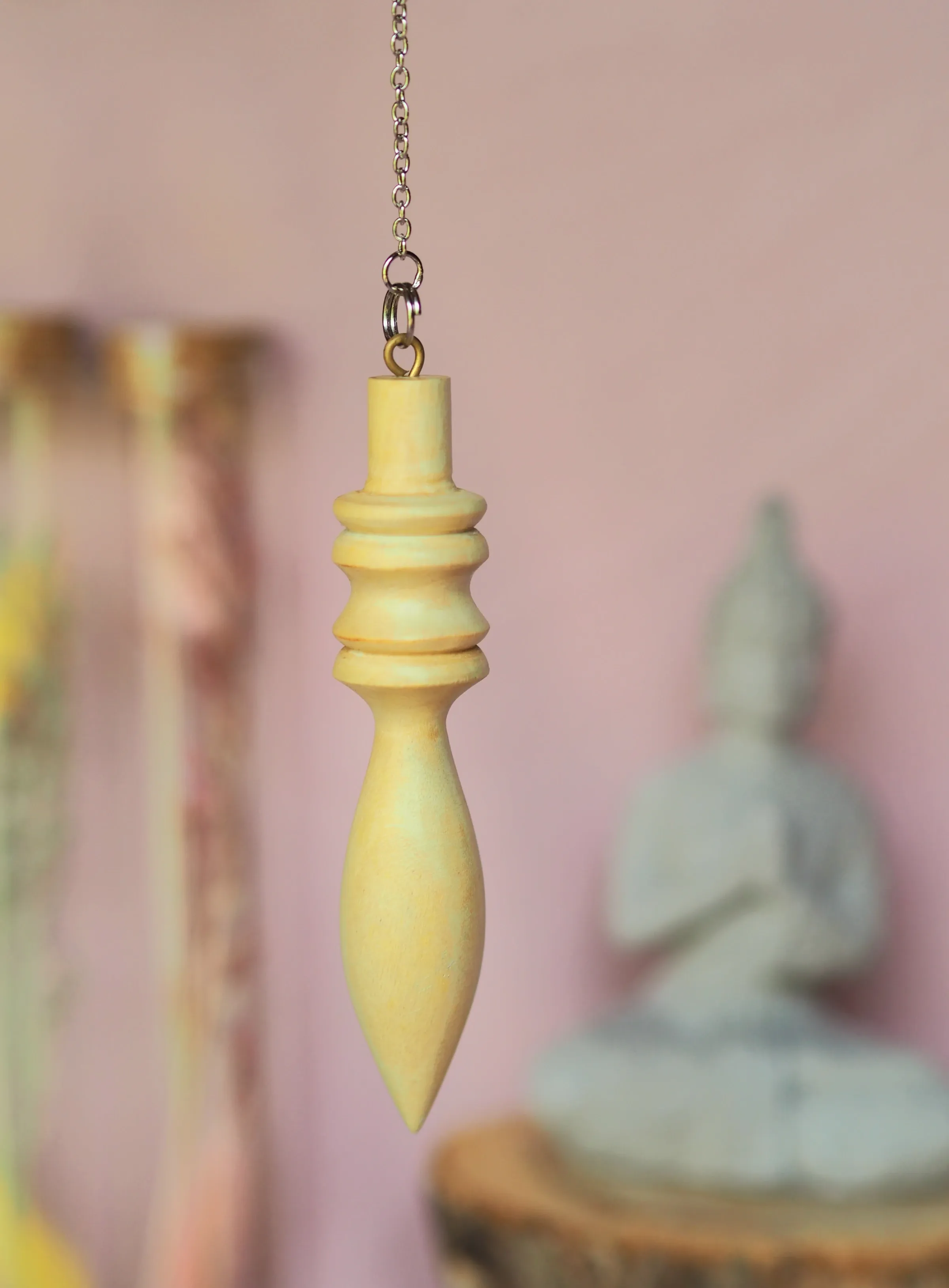 Pendule en bois clair, cerclé de deux anneaux et effilé en pointe, suspendu à une chaînette devant un mur rose