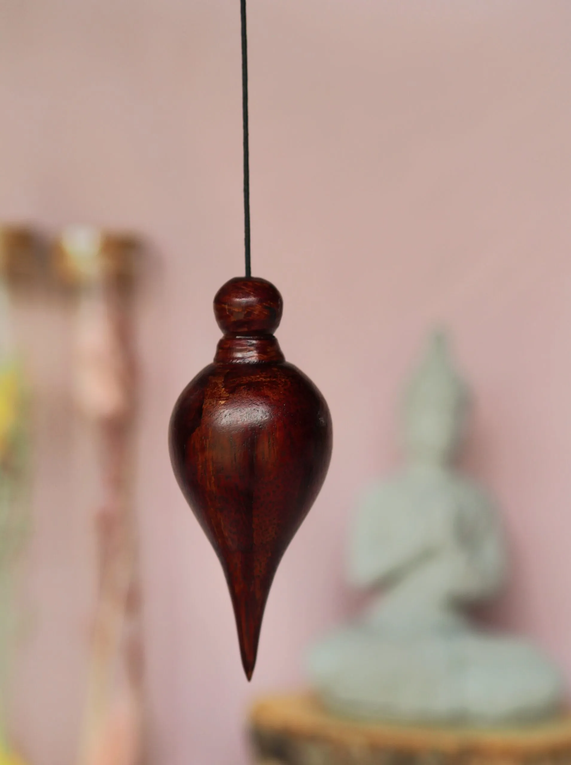 Pendule en bois brun-rouge en forme de goutte d’eau, suspendu à un cordon noir, devant un mur rose et une statuette de Bouddha