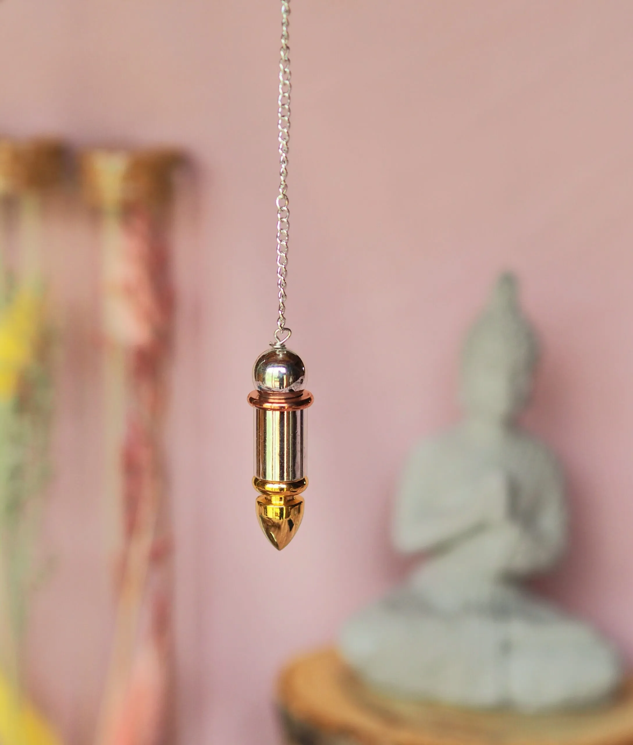 Pendule en acier en forme d’obus, tête et corps argentés, pointe dorée, suspendu à une chaînette devant un mur rose