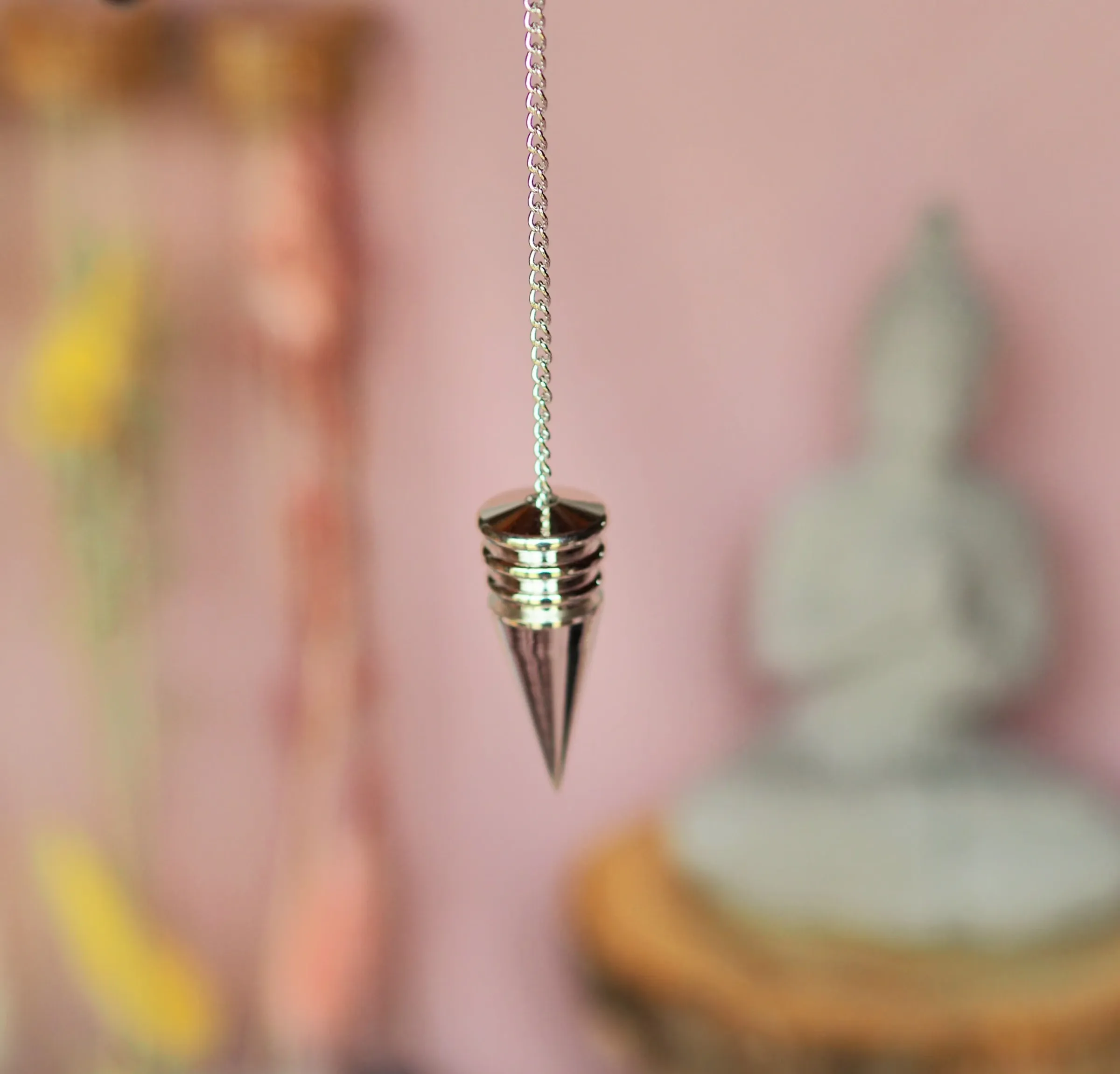Pendule argenté à tête cerclée d’anneaux et long cône effilé, suspendu à une chaînette devant un mur rose