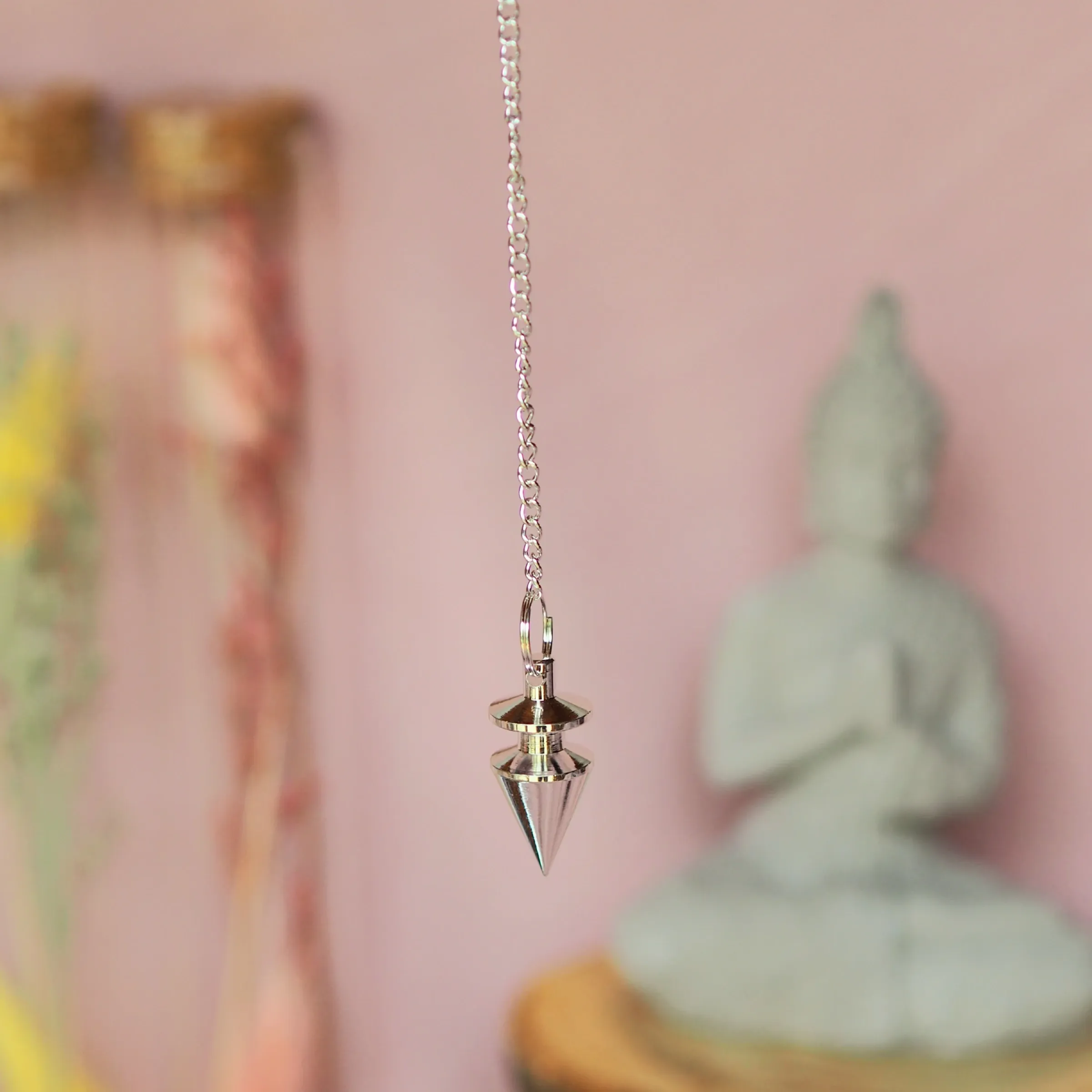Petit pendule argenté, un disque au-dessus d’un cône pointu, suspendu à une chaînette devant une statuette de Bouddha