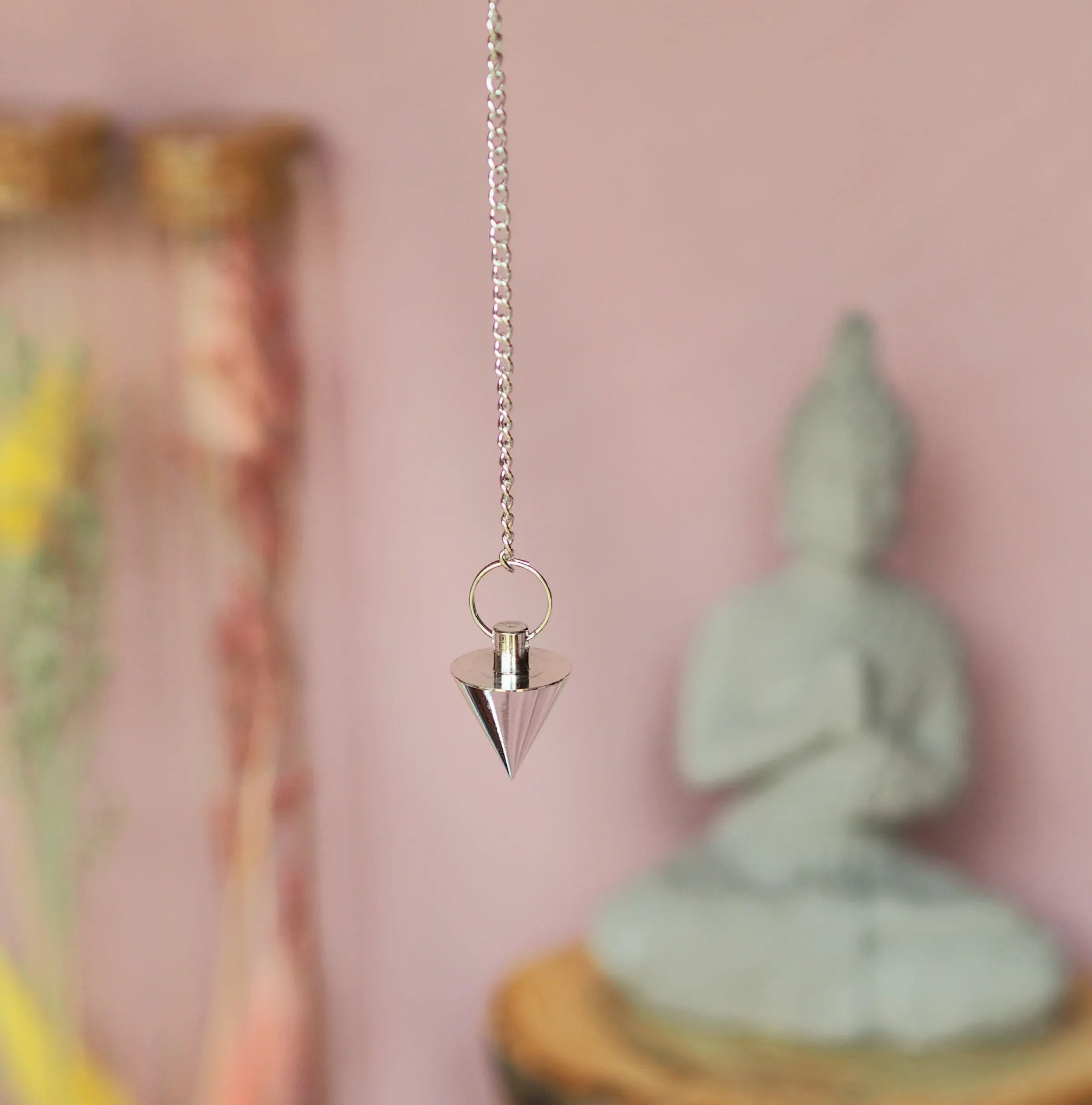 Petit pendule conique en acier argenté, suspendu à une chaînette fine devant une statuette de Bouddha