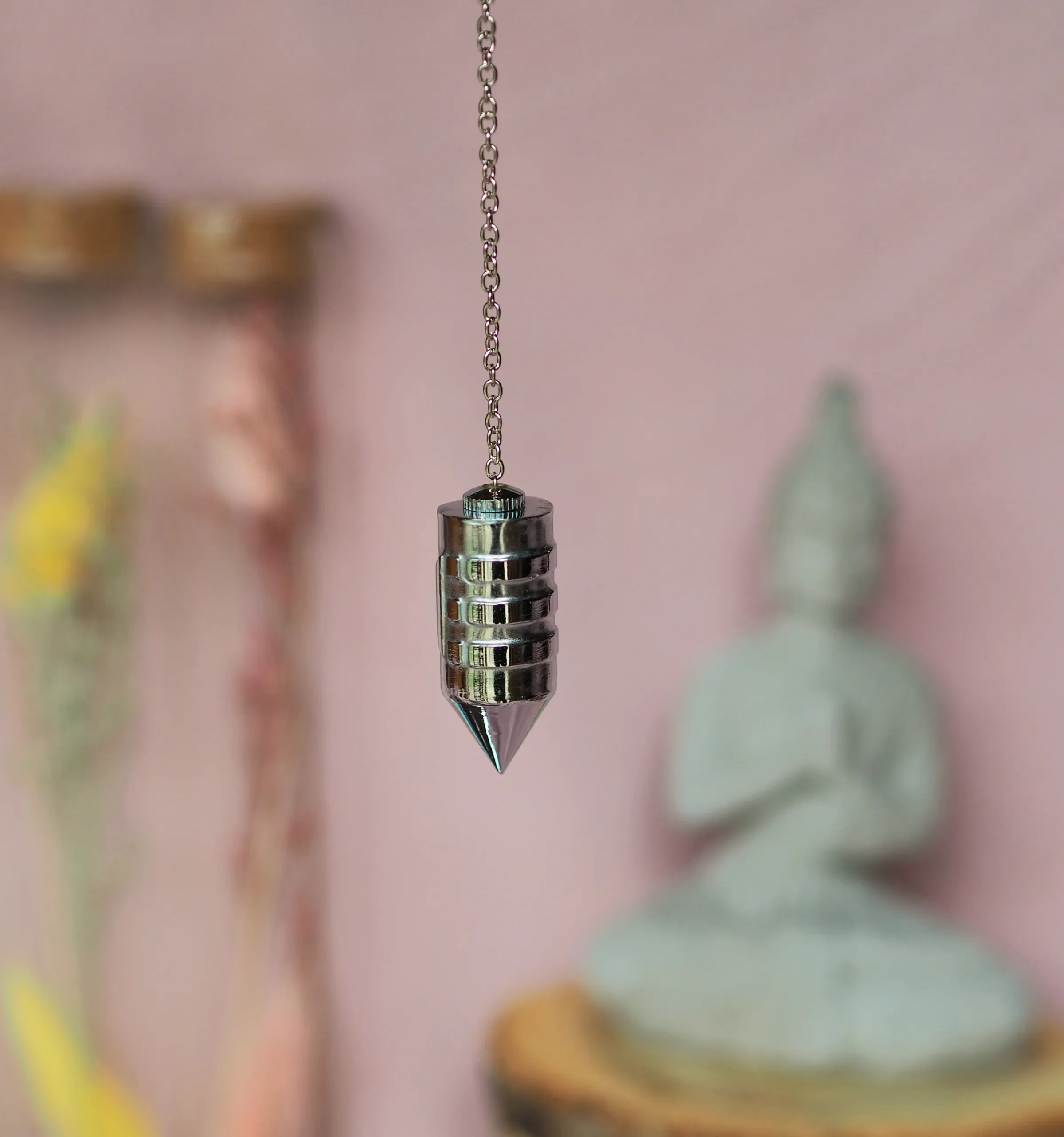 Pendule chromé au corps cylindrique cerclé d’anneaux et à la pointe conique, suspendu à une chaînette
