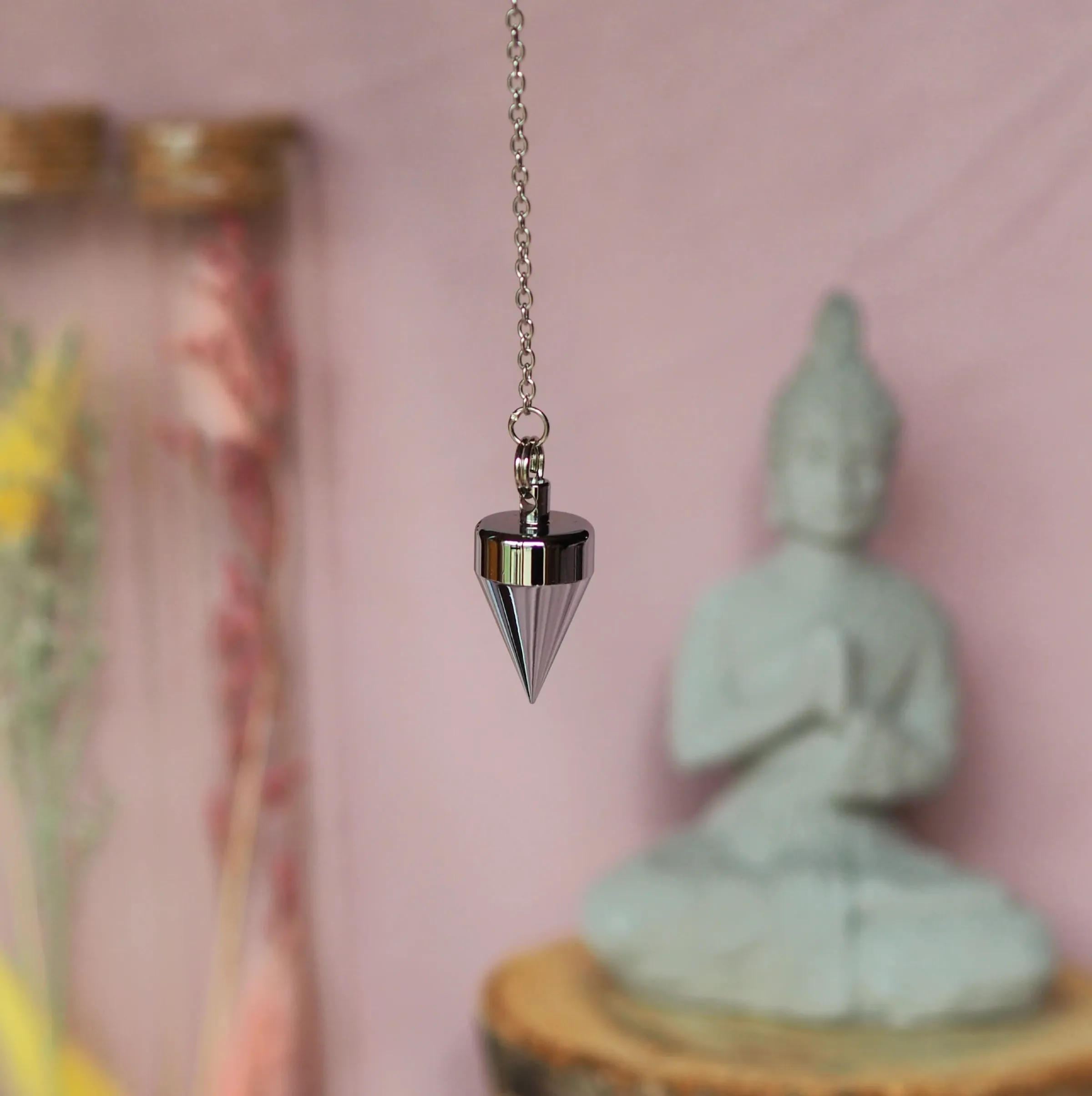 Pendule chromé à tête cylindrique et cône pointu, suspendu à une chaînette devant une statuette de Bouddha