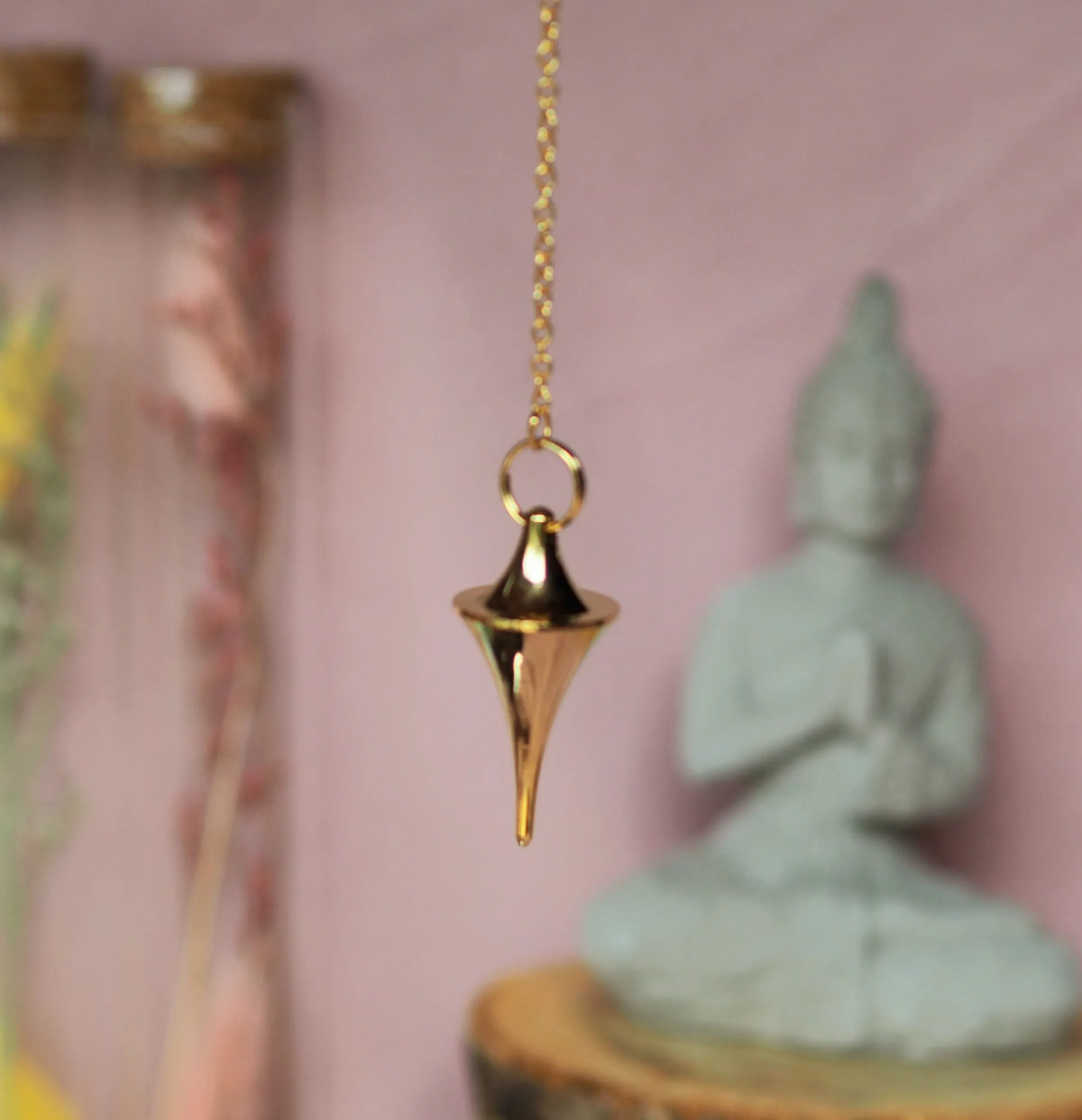 Pendule doré en forme de toupie effilée, suspendu à une chaînette dorée devant une statuette de Bouddha